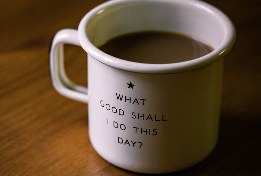 White coffee cup with a star on the side and the saying What good shall I do today?, filled with coffee and sitting on a wooden table