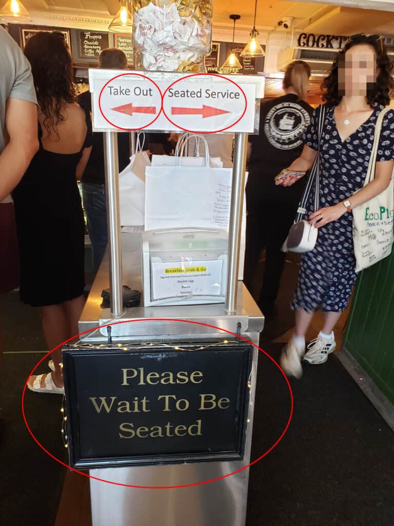 Main Streets Market and Cafe entry way close up of the cart bisecting the entryway, with two small signs at shoulder height. The on one the left says take out. The right one says seated service. A third sign at hip height reads "please wait to be served." All three signs are highlighted with red ovals.