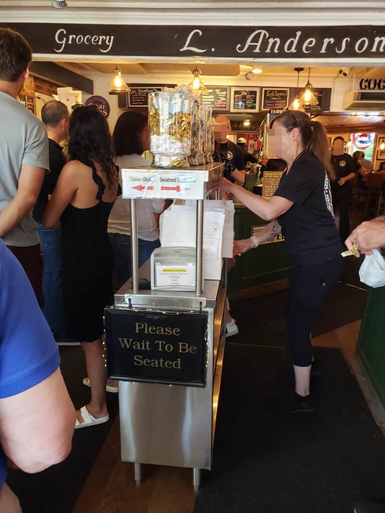 Scene entering Main Streets Market and Cafe in Concord, Massachusetts, with a line of people to the left of a two-tier, stainless-steel cart, and a server on the right side. Black carpet runners are on either side of the cart, leading to the take-out counter.