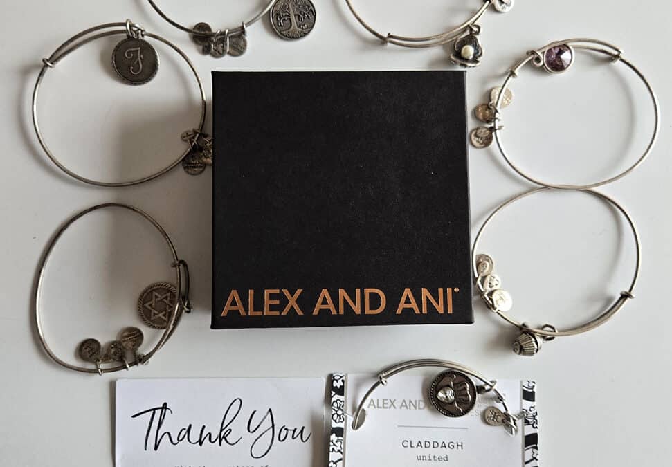 Black 3 1/2" square jewelry box with Alex and Ani on it in gold capital letters, surrounded by silver wire bracelets bearing charms featuring a script letter F, a Jewish star, a Claddagh, a purple gemstone, a weathered silver flower with a pearl in the middle, and a tree of life