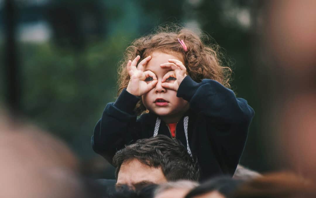 a 4 year old girl perched on her father's shoulders wearing a navy blue sweatshirt, an orange barrette in her curly brown hair, and using her thumb and pointer fingers to encircle both eyes like she is looking through binoculars